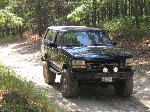 1995 Ford Bronco - This lifted black Ford Bronco is driving on a dirt road surrounded by trees.
