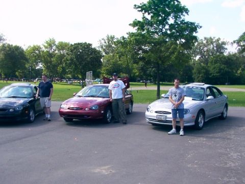 1998 Ford Taurus - Three Ford Taurus sedans are parked in a lot with three men standing next to them.