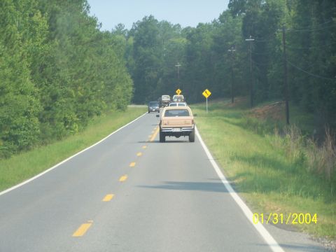 1979 Ford F-150 - A line of vehicles travels down a rural highway bordered by trees.