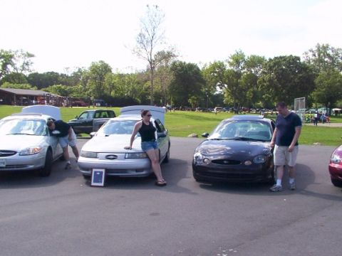 1998 Ford Taurus - Two Ford Tauruses are parked at an outdoor event, one with its hood up and a woman posing.