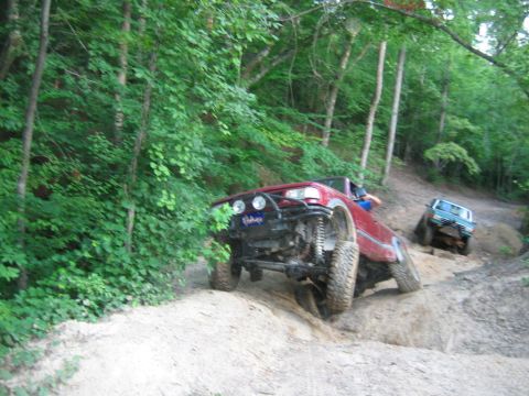 1992 Ford Mustang - Two trucks traverse a wooded off-road trail with steep inclines and loose dirt.
