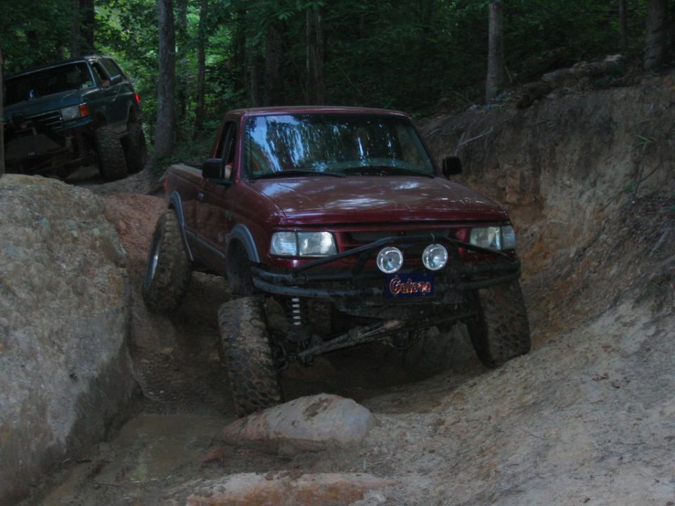 Red pickup truck with off-road tires navigating a muddy trail, green SUV visible in background