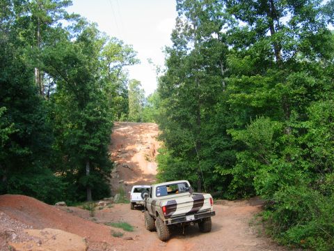 1992 Ford Mustang - Two trucks navigate a challenging off-road trail surrounded by dense green trees.