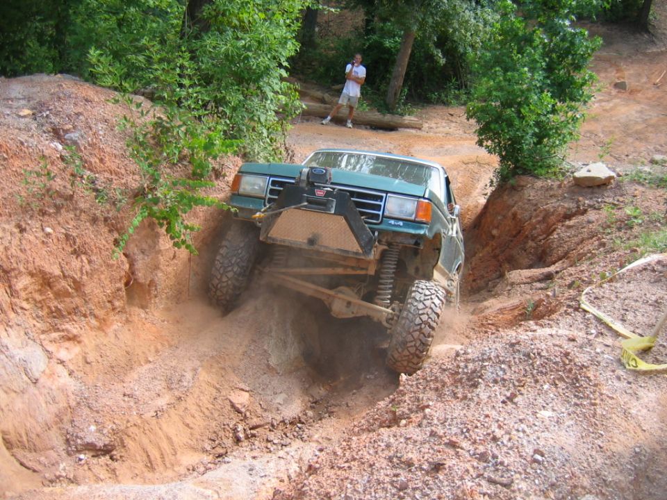 Green pickup truck with winch on front bumper going down a dirt trail with steep sides. Person standing in background
