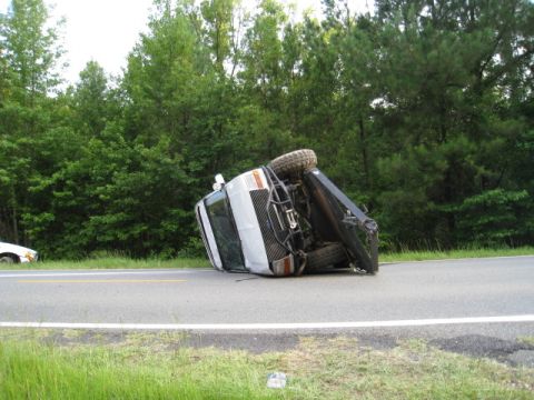 1979 Ford F-150 - A white pickup truck lies on its side on the side of a road, with trees in the background.