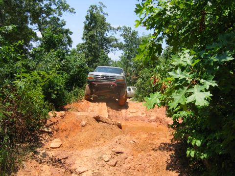 1992 Ford Mustang - A silver pickup truck drives through a challenging muddy off-road course with trees lining the path.