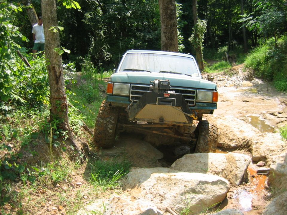 Green SUV with large tires navigating a rocky, muddy trail in the woods. Winch mounted on front bumper