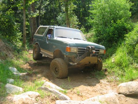 1992 Ford Mustang - A green SUV with off-road modifications drives through a challenging, rocky terrain surrounded by trees.