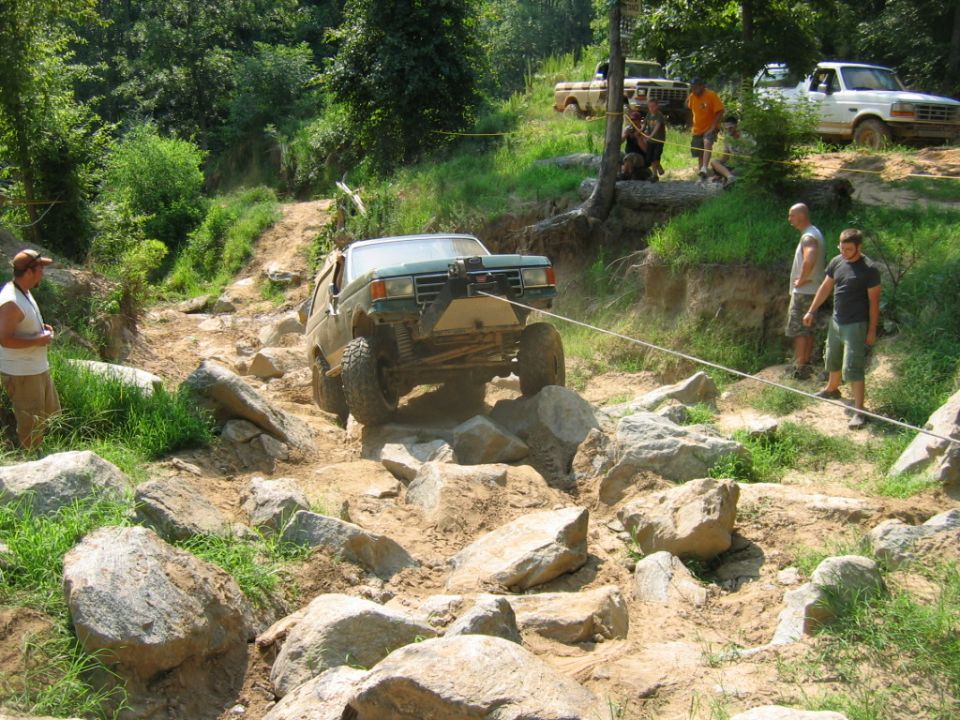 Green off-road truck with winch cable on rocky trail; other trucks and people in background