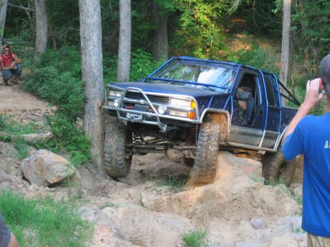 1992 Ford Mustang - A modified blue pickup truck navigates a challenging off-road trail with large tires and a protective roll cage.