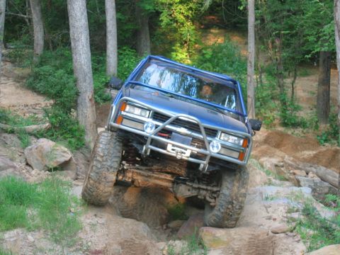 1992 Ford Mustang - A blue off-road vehicle tackles a challenging dirt and rock path in a wooded area.