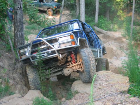 1992 Ford Mustang - A heavily modified blue truck with a roll cage tackles a challenging off-road course, with another vehicle visible in the background.