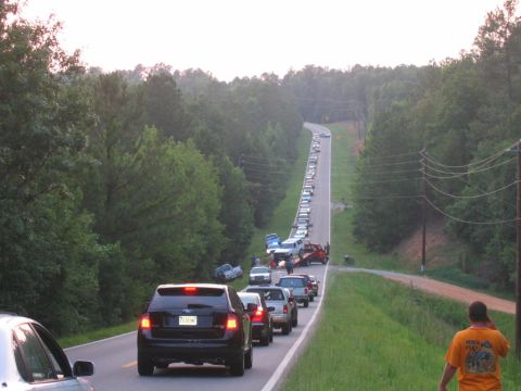 1992 Ford Mustang - A significant traffic jam stretches along a tree-lined rural road, with vehicles queued for a considerable distance.