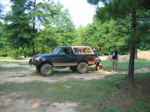 1992 Ford Mustang - A dark blue Ford Bronco is parked outdoors, with two people near its rear and a third person standing nearby.