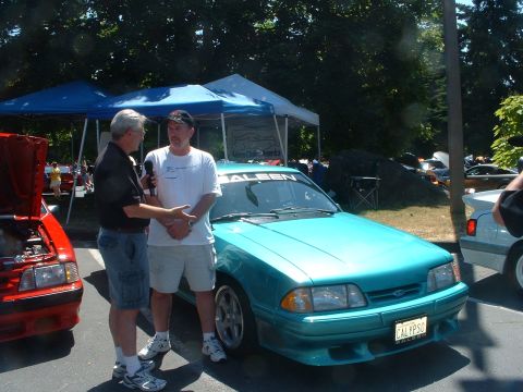2000 Ford Crown Victoria - A teal Saleen Mustang is shown with two men talking in front of it, with another red car visible to the left.