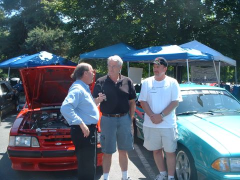 2000 Ford Crown Victoria - Three men converse at an outdoor car show, with a red car and a teal car visible.