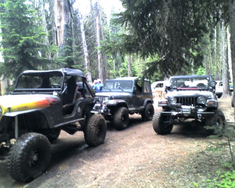 2005 Jeep Wrangler - Three modified Jeep Wranglers are parked on a dirt path in a forest. The Jeep in the foreground has a yellow hood with flame decals and lar…