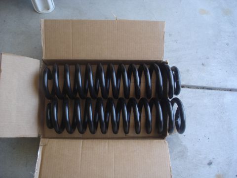1987 Ford Bronco - Two black coil springs are shown lying side-by-side inside an open cardboard box. The springs are coiled tightly and appear to be new.