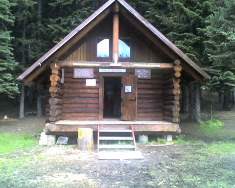 2005 Jeep Wrangler - A rustic log cabin with a wooden door and windows is surrounded by tall pine trees. A wooden sign above the door reads "CAMP MIKE URICH".