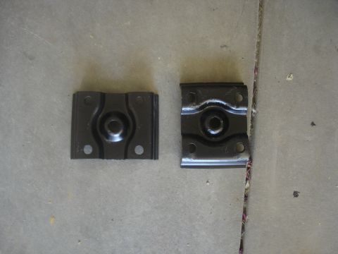 1987 Ford Bronco - Two black metal brackets with central circular indentations and four mounting holes are shown on a concrete surface.