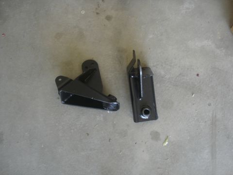 1987 Ford Bronco - Two black metal brackets are shown lying on a concrete surface. One bracket is triangular and open on one side, while the other is rectangu…