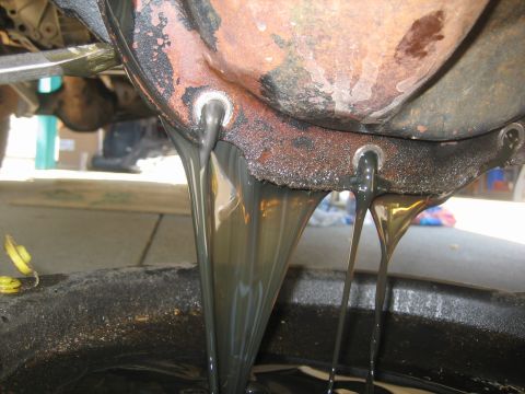 1995 Ford Bronco - Dark, used oil is draining from the differential housing of a vehicle into a black oil drain pan. The differential housing appears rusty an…