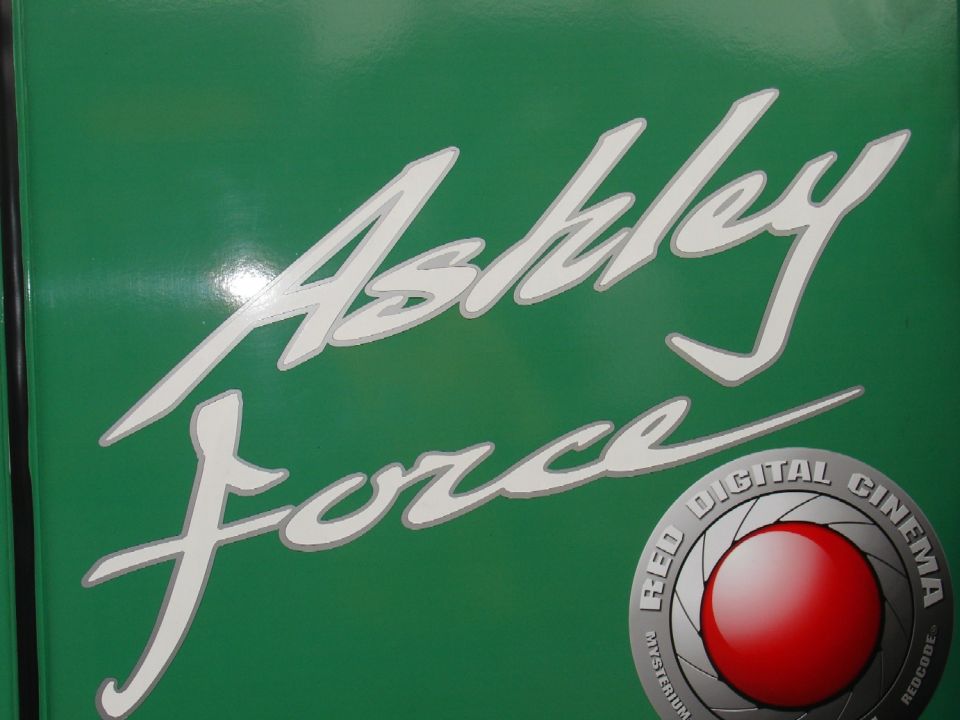 White script text "Ashley Force" on a green background with a red lens logo.