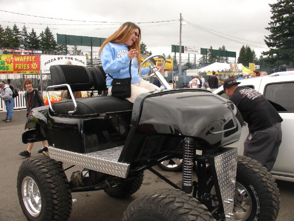 Woman in blue sweatshirt laughing in a black, lifted golf cart with large tires.