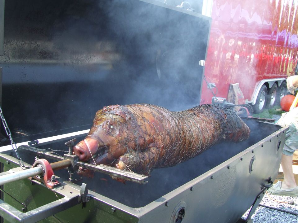 Whole pig roasting on a spit with smoke, red trailer in background