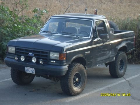 1990 Ford Bronco - A dark blue Ford Bronco is parked on asphalt with vegetation in the background. The vehicle has a spare tire mounted on the rear and afterm…