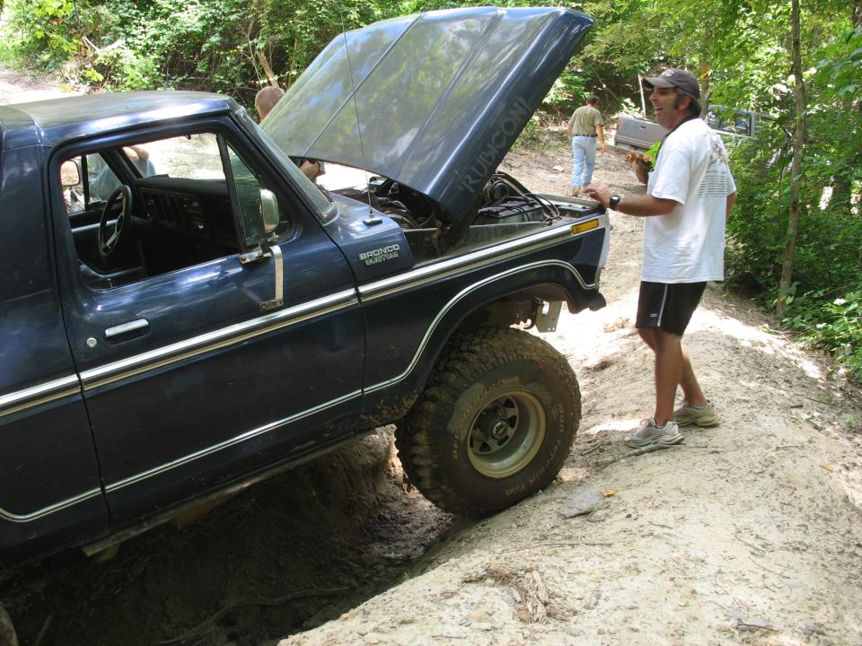 Man at the open hood of a blue Ford Bronco on a rutted dirt trail