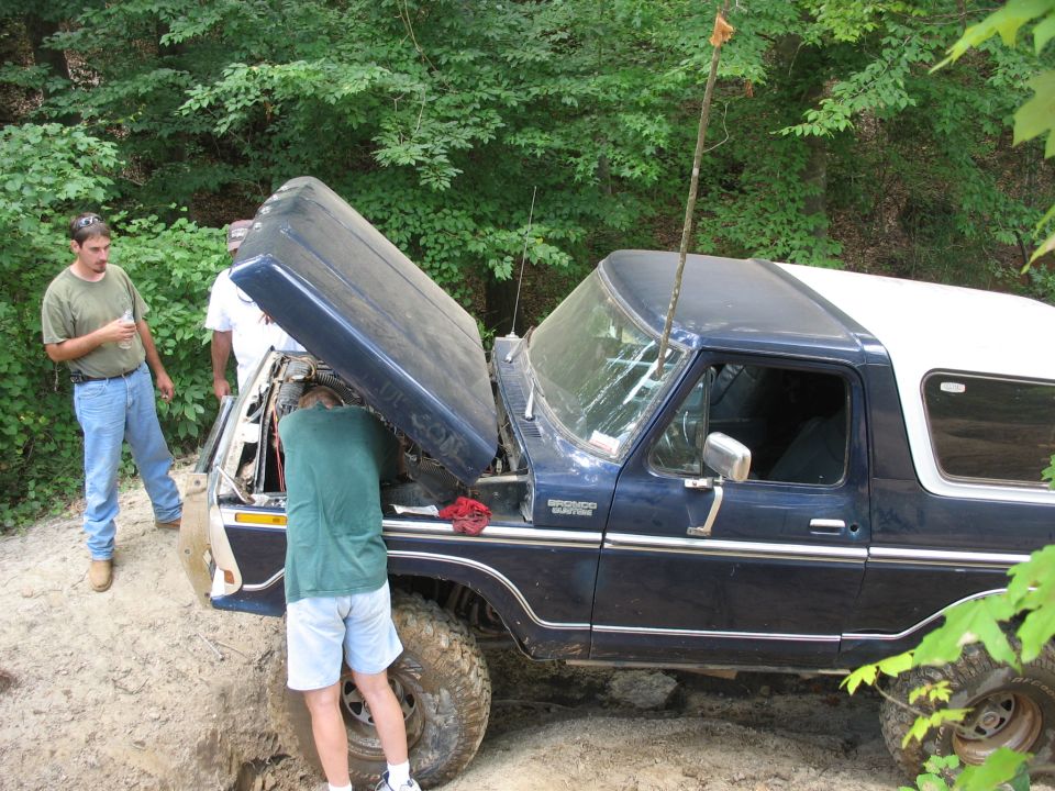 People working on a tilted blue Ford Bronco on a rocky off-road trail
