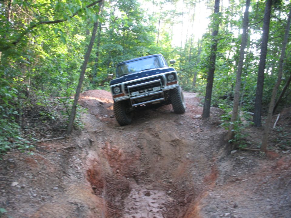 Ford Bronco descending toward a deep red-dirt rut on a wooded trail