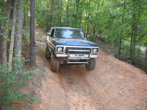 1978 Ford Bronco - A blue 1978 Ford Bronco navigates a challenging off-road trail through a wooded area.