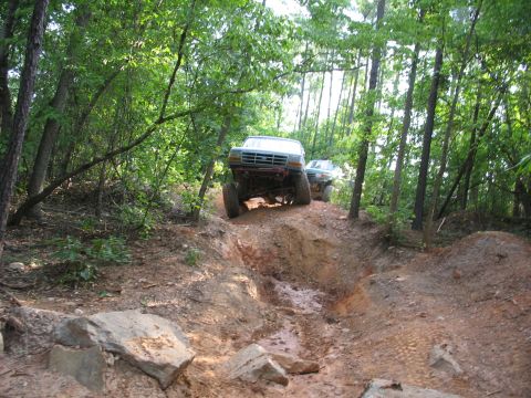 1978 Ford Bronco - Two off-road vehicles navigate a challenging dirt path through a dense forest.