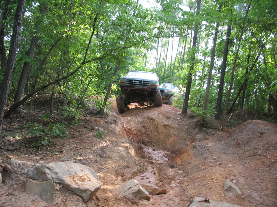 Two lifted trucks driving up a muddy, wooded trail. The lead truck is a light blue SUV