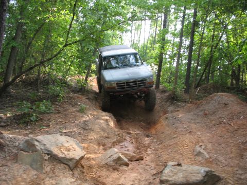 1978 Ford Bronco - A light blue SUV navigates a challenging off-road trail through a wooded area.