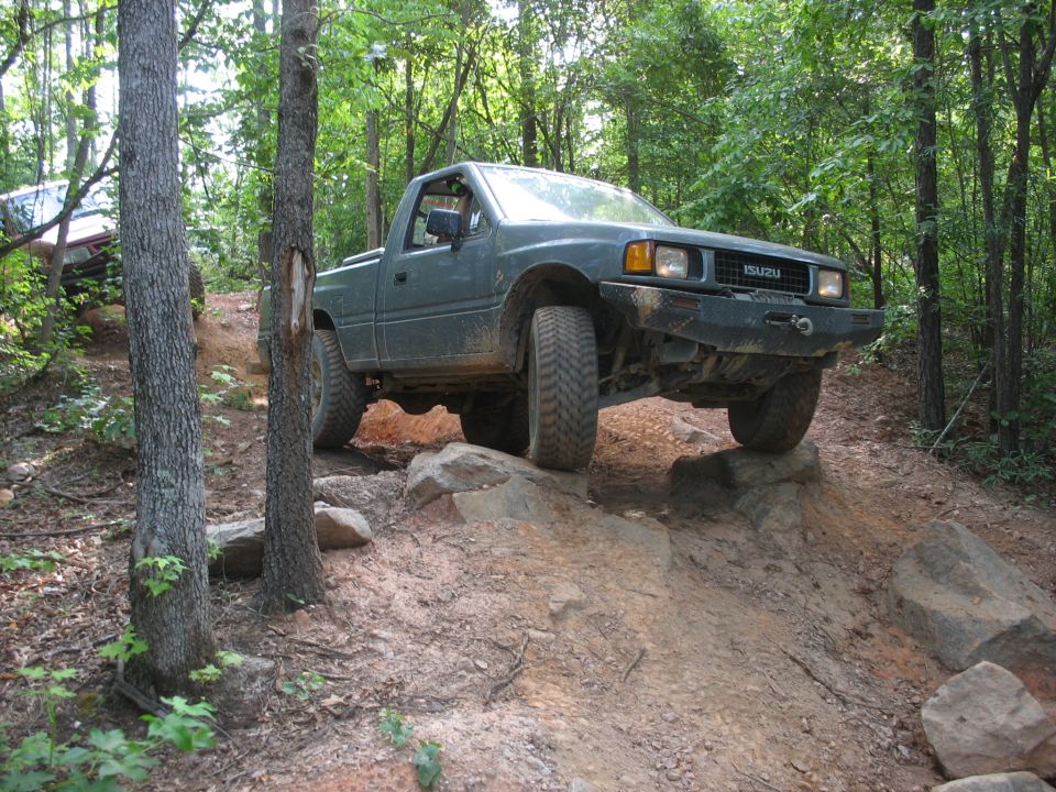 Green Isuzu pickup truck climbing over rocks on a dirt trail in the woods. Another vehicle is visible behind it