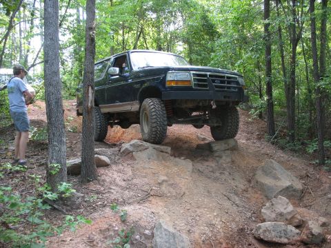 1978 Ford Bronco - A dark SUV with off-road modifications is shown traversing a challenging rocky trail in a wooded area.