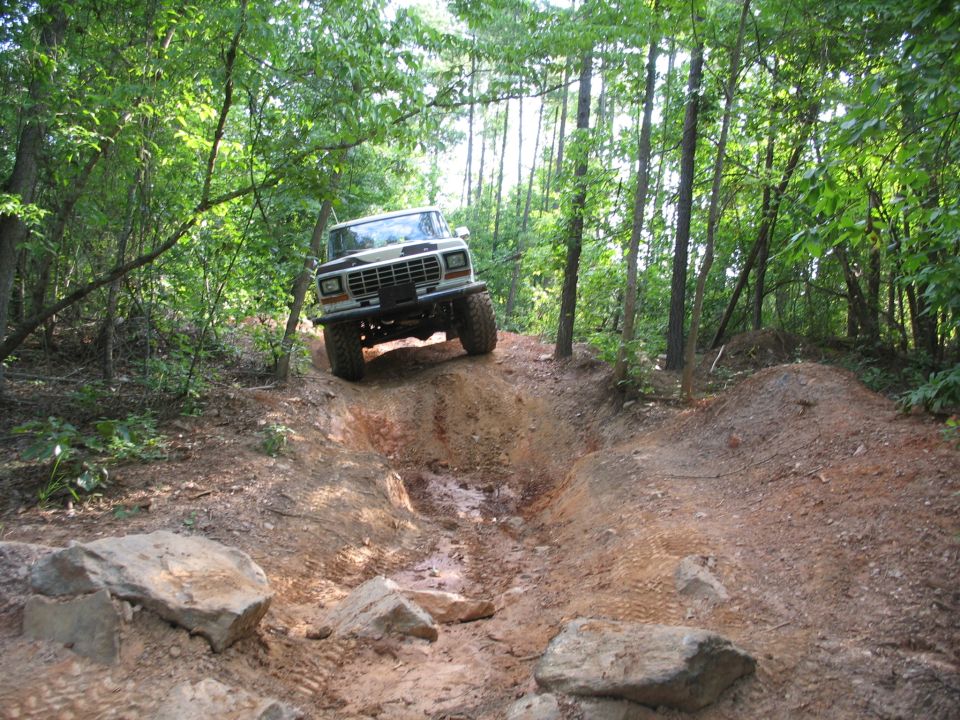 White 1978 Ford Bronco driving up a muddy, rocky hill in a forest