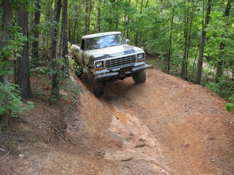 1978 Ford Bronco - A tan and camouflage pickup truck navigates a muddy, wooded trail.