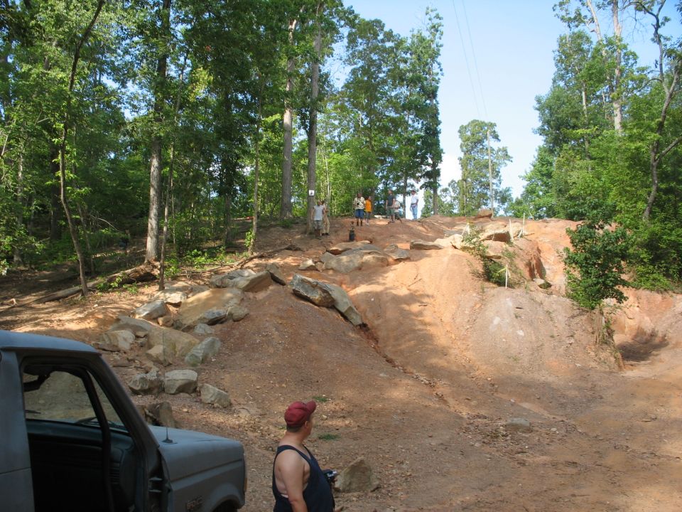 Partial view of a truck parked on dirt near an off-road trail with rocks and trees; several people are visible on the trail