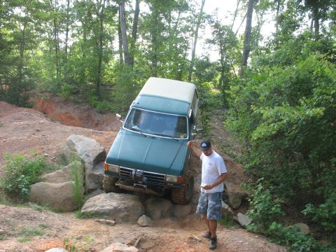 1978 Ford Bronco - A man watches as an off-road vehicle carefully maneuvers over a rocky obstacle on a wooded trail.