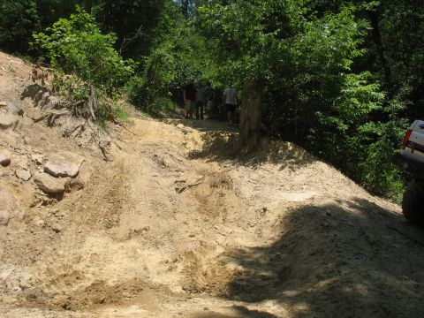 1978 Ford Bronco - A white SUV is partially visible on the right side of a muddy, rutted dirt trail with several people standing in the background.