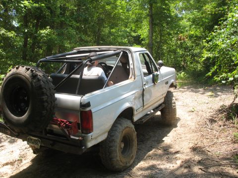 1978 Ford Bronco - White SUV with a roll cage and spare tire drives on a dirt path through a wooded area.