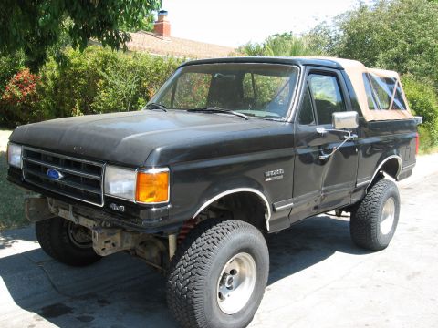 1989 Ford Bronco - Front three-quarter view of a lifted black late-1980s Ford Bronco with the hardtop replaced by a tan soft top, parked on a residential driv…