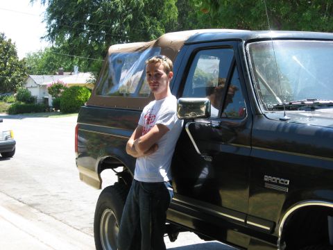 1989 Ford Bronco - A person in a white graphic tee leans arms-crossed against the driver's door of a black late-1980s Ford Bronco fitted with a tan soft top,…