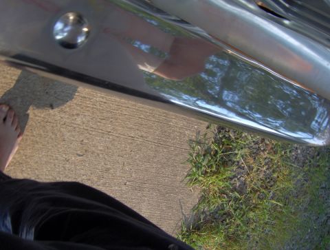 1978 Ford Bronco - A close-up of a chrome bumper, reflecting trees and a person's leg, with a concrete and grass foreground.