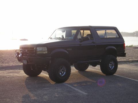 1995 Ford Bronco - This black Ford Bronco SUV, likely from the early to mid-1990s, is modified for off-road use and parked near a beach at sunset.