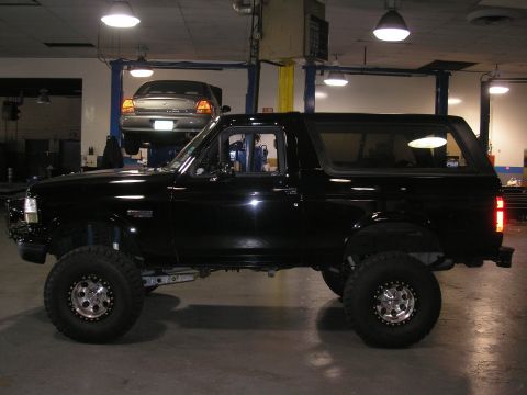 1995 Ford Bronco - A black Ford Bronco is parked in a garage, with another car lifted on a car hoist in the background.